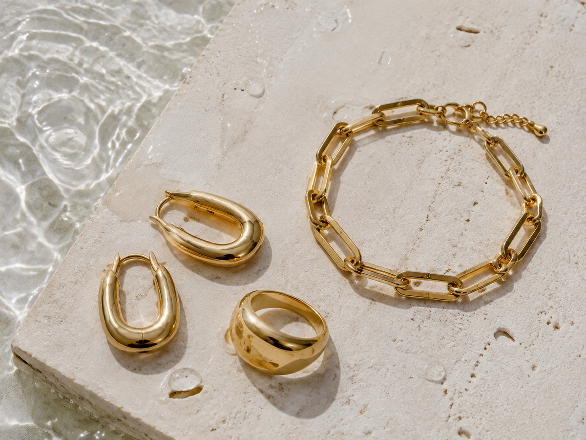 Gold-tone PVD stainless steel jewelry beside water on pale stone